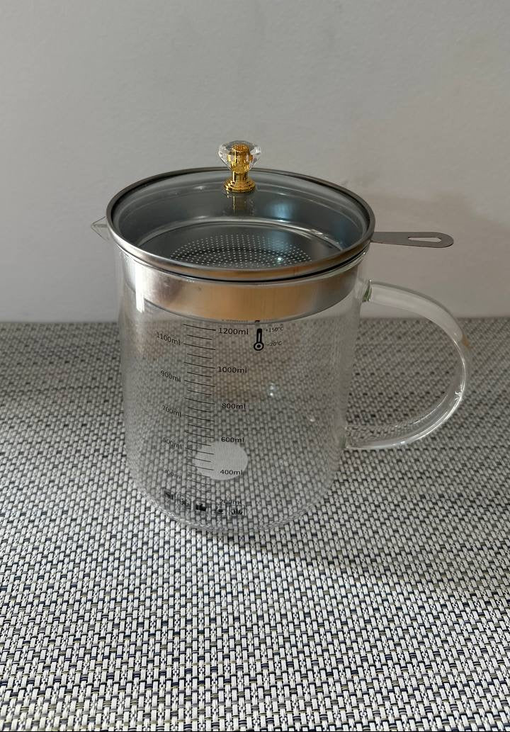Clear glass teapot with metal lid and filter on a textured surface.