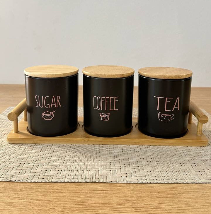 Three black canisters labeled 'Sugar', 'Coffee', and 'Tea' on a wooden tray with a white background.